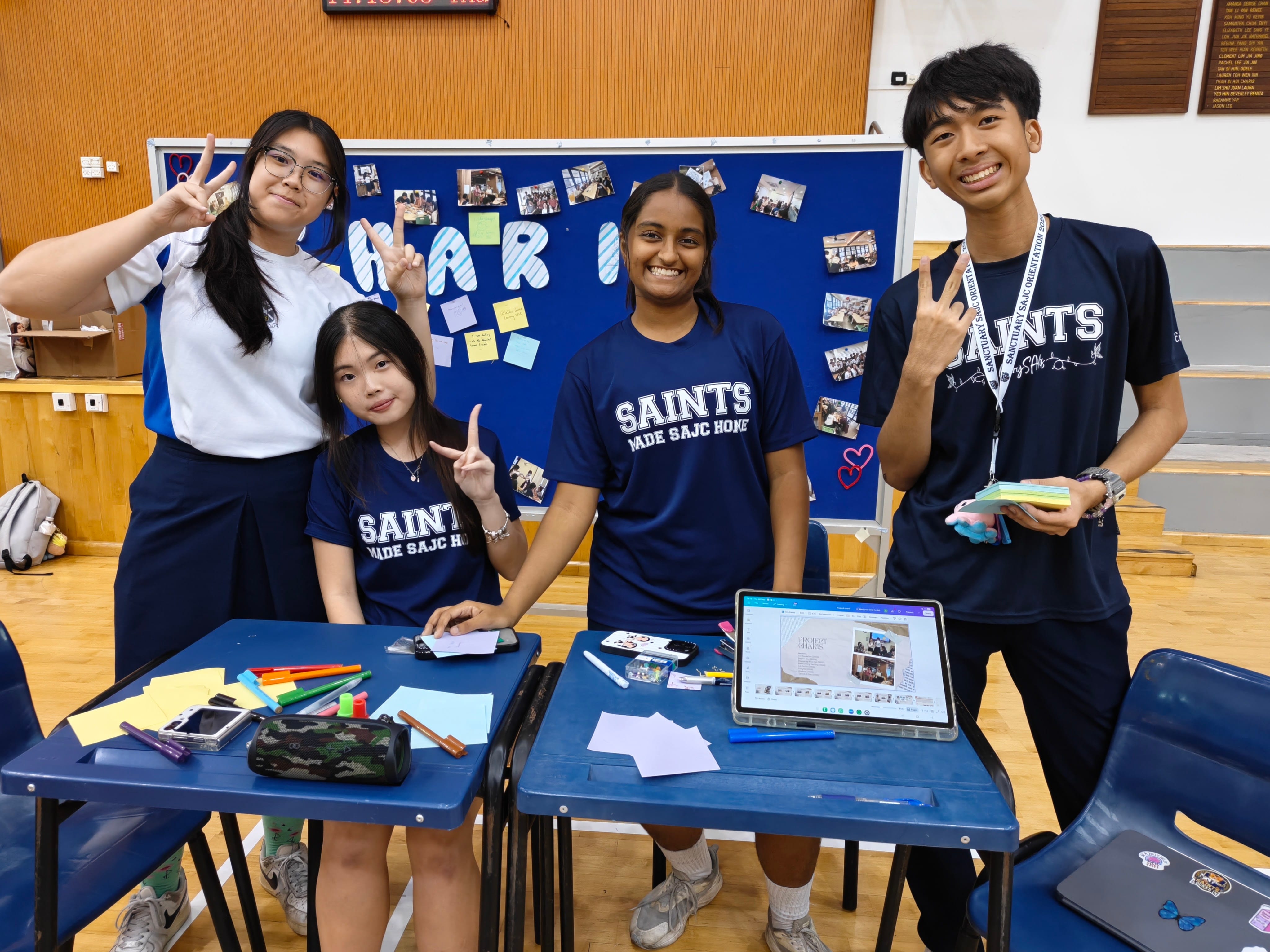 Four smiling students making peace signs behind a classroom desk setup at a charity exhibition booth with sticky notes and photo displays.